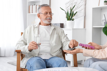 Senior man with glass of water taking pill from caregiver in nursing home