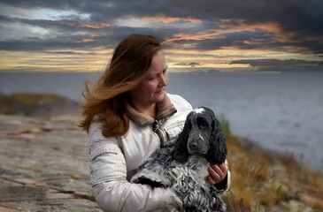 Portrait of a woman and an English Cocker Spaniel puppy. She sits on the rocky shore of the island and holds a dog in her hands. In the background there is a lake and a beautiful sunset.