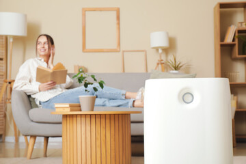 Modern humidifier on floor against young woman sitting on sofa in living room