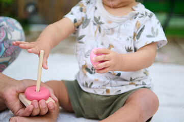 Close-up with focus on hands of a toddler stacking round circles of educational developmental wooden pyramid, playing with his mother supervising him, sitting together on the linen blanket outdoors