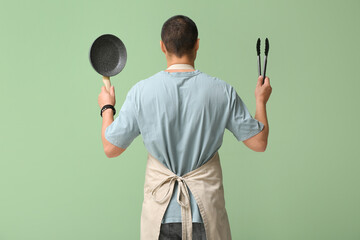 Young man in apron with frying pan and tongs on green background, back view