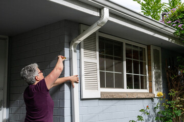 Middle aged white woman using a screwdriver to re-install a rain gutter downspout after cleaning it out, homeowner maintenance
