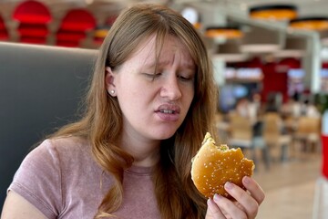 Sad depressed young woman eating burger in fast food restaurant. Eating disorder concept. 