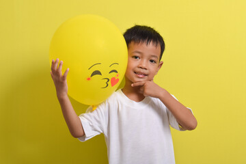 Kiss emoji. Cute Asian boy holding kiss emoticon balloon isolated on yellow background