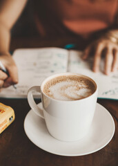 cup of coffee with milk with a female hands writing on a notebook at the background