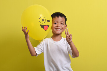 Tongue emoji. Cute Asian boy holding tongue out emoticon balloon isolated on yellow background. Teasing emoji with closed eyes