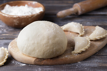 Dough for dumplings on a wooden cutting board, ready-made dumplings, a bowl of flour and a rolling pin for rolling out dough on a wooden table