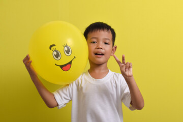 Smile emoji. Cute Asian boy holding happy emoticon balloon isolated on yellow background