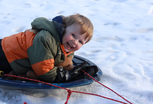 Smiling child sledding on snow with a teddy bear