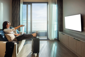 Mid aged asian businesswoman relax in home or hotel resting after travelling using laptop. Woman choosing movie on internet streaming service with remote control in hand. White screen LED TV isolated.