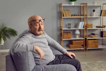 Fototapeta premium Portrait of a senior man at home. Retired old bald man in a gray sweater, with glasses on his forehead, sitting on the sofa in the living room and looking at the camera with a serious face expression