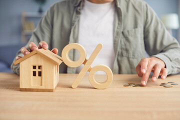 Wooden percent symbol and house model on table with crop young man counting coins in background....