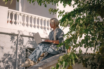 Fototapeta premium A woman is sitting on a white railing with a laptop in front of her. She is wearing a blue jacket and jeans. Concept of relaxation and leisure.