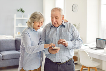 Portrait of happy smiling senior couple standing in living room at home and using mobile phone together for online shopping, texting or social media. People in retirement and technology concept.