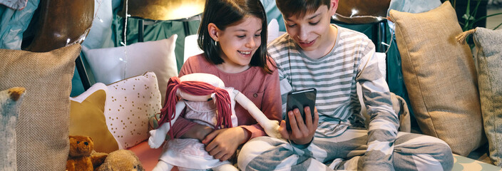 Banner of happy children laughing together while watching video on cell phone sitting on shelter tent at home. Smiling little girl embracing rag doll next to boy using mobile in a cozy diy teepee.