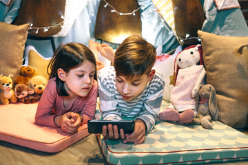 Children watching unsafe content on cellphone without parental supervision at home. Girl and boy lying together while chatting with strangers on social network app. Screen addiction dangers concept.