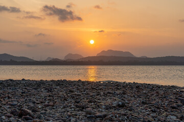 The sunset over the calm sea with the reflection reflecting the beauty of nature, the view of the mountains below and the beach with many beautiful rocks, in the atmosphere of brown and yellow light, 