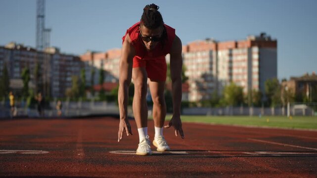 Slow motion. A male athlete in sunglasses and dreadlocks on his head takes the crouch start position, while standing on the running track of a city stadium outdoors on a warm autumn day. The man