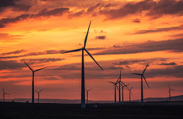 Fototapeta premium Multiple wind turbines silhouetted against a vibrant sunset sky. The turbines stand tall against the backdrop of orange and red clouds.
