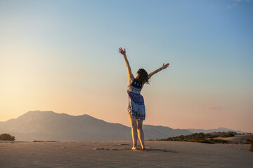 Obraz premium Beautiful woman with hands up looking on the sunset standing on the sand against mountain and sky