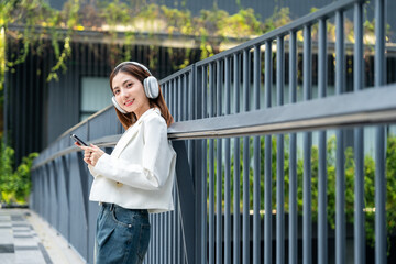 Young asian woman using smartphone while walking in the urban city. Happy Beautiful asian female with headphone listen to the music by cell phone standing at office building city. Lifestyle people