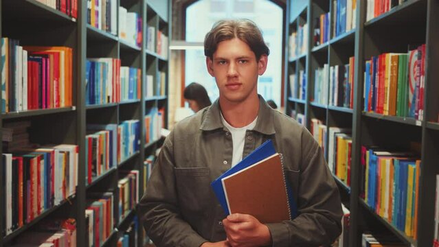Young man in gray jacket holding notebooks in library aisle, looking directly at camera. Colorful books and another student studying. Concept of academic focus and scholarly dedication