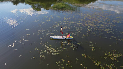Two children enjoying summer holiday and sunny day, paddling stand up board on a calm lake water, aerial shot. Carefree childhood concept.