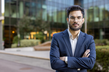 Confident young businessman standing outdoor with arms crossed in front of modern office building....