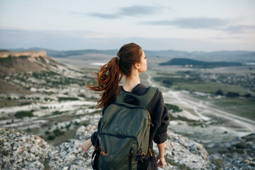 Woman standing on mountain peak gazing at valley below with backpack on sunny day
