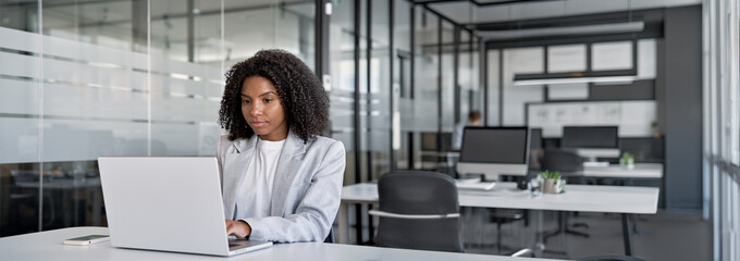 Young Latin businesswoman using computer, working in modern office. Focused African American woman doing online business data market analysis, planning tech strategy at laptop pc. Banner, copy space