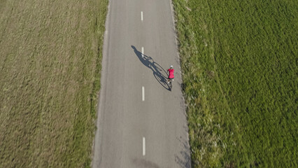 Rear view of a female racing cyclist riding on an empty asphalt road with a large shadow of body and bike, aerial tracking drone shot.