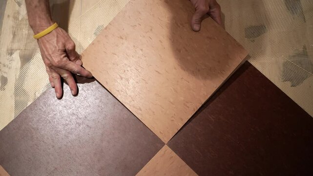 With glue on the floor, two hands hold a VCT tile and carefully move it into place, pressing it down into the glue.