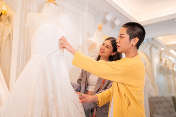 Naklejka premium Asian bride is smiling while choosing wedding dress in modern wedding salon, Attractive designer girl using tape meter fitting on wedding dress at wedding studio.