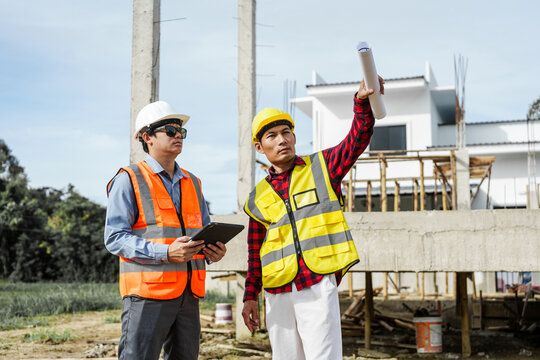 Two male construction workers, including an Asian engineer, meticulously review structural plans and design concrete columns and beams for a new residential build.