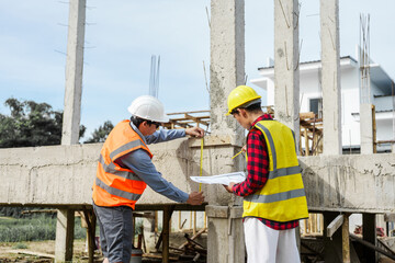 Two male construction workers, including an Asian engineer, meticulously review structural plans...