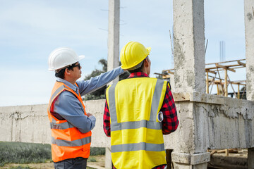 Two male construction workers, including an Asian engineer, meticulously review structural plans and design concrete columns and beams for a new residential build.