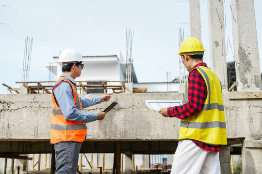 Two male construction workers, including an Asian engineer, meticulously review structural plans and design concrete columns and beams for a new residential build.