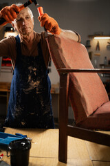 Obraz premium An elderly man in gloves and a blue apron begins to restore an old chair in a workshop. holds a hammer removes old fabric. concept second life of old furniture. Copy space.