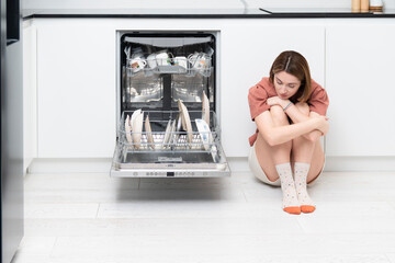 Tired and bored woman putting dirty plates in dishwasher machine in the kitchen. Household and...