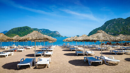 Tranquil beach setting in Marmaris with straw umbrellas, white sun loungers on sandy shore. Calm blue sea, picturesque mountains under clear sky. Welcome to Marmaris, Turkey concept image © IgorZh