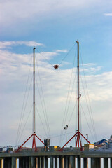 Two towering masts support a thrilling suspended ride. The waterfront setting suggests an amusement park or carnival atmosphere, with hints of urban structures in the background.