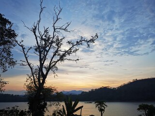 Serene lakeside sunset: silhouetted trees frame golden sky over mountains. Lone boat on calm waters embodies tranquility, natural beauty, and peaceful solitude.