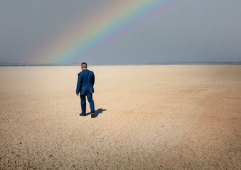 A man wearing  a blue suit walking on a dry lake bed in the desert with a rainbow