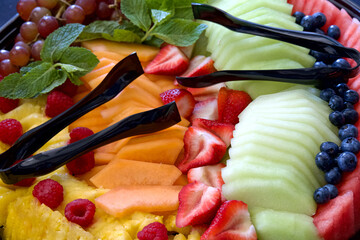 Top view close up of fruit tray with sliced melons, straberries, watermelon, cantaloupe, grapes and pineapple. Tongs laying on top for serving.