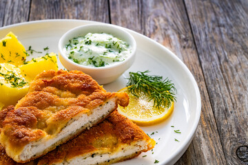 Crispy breaded seared chicken cutlet with boiled potatoes and cucumbers in cream served on white plate on wooden table