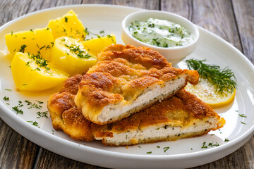Crispy breaded seared chicken cutlet with boiled potatoes and cucumbers in cream served on white plate on wooden table