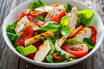 Caesar style salad - delicious grilled chicken breast slices and fresh vegetables and seasonings served in white bowl on wooden table
