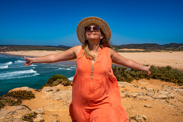 Beautiful woman in fashionable orange dress at viewpoint on cliff near sunny and sandy beach...