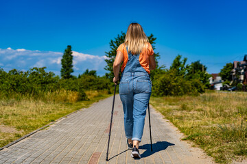 Nordic walking -  mid-adult beautiful woman in jeans dungarees exercising on footpath using Nordic...