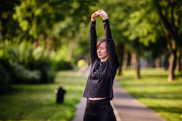 A young woman in black sportswear stretching her arms upwards on a park path, preparing for a run....